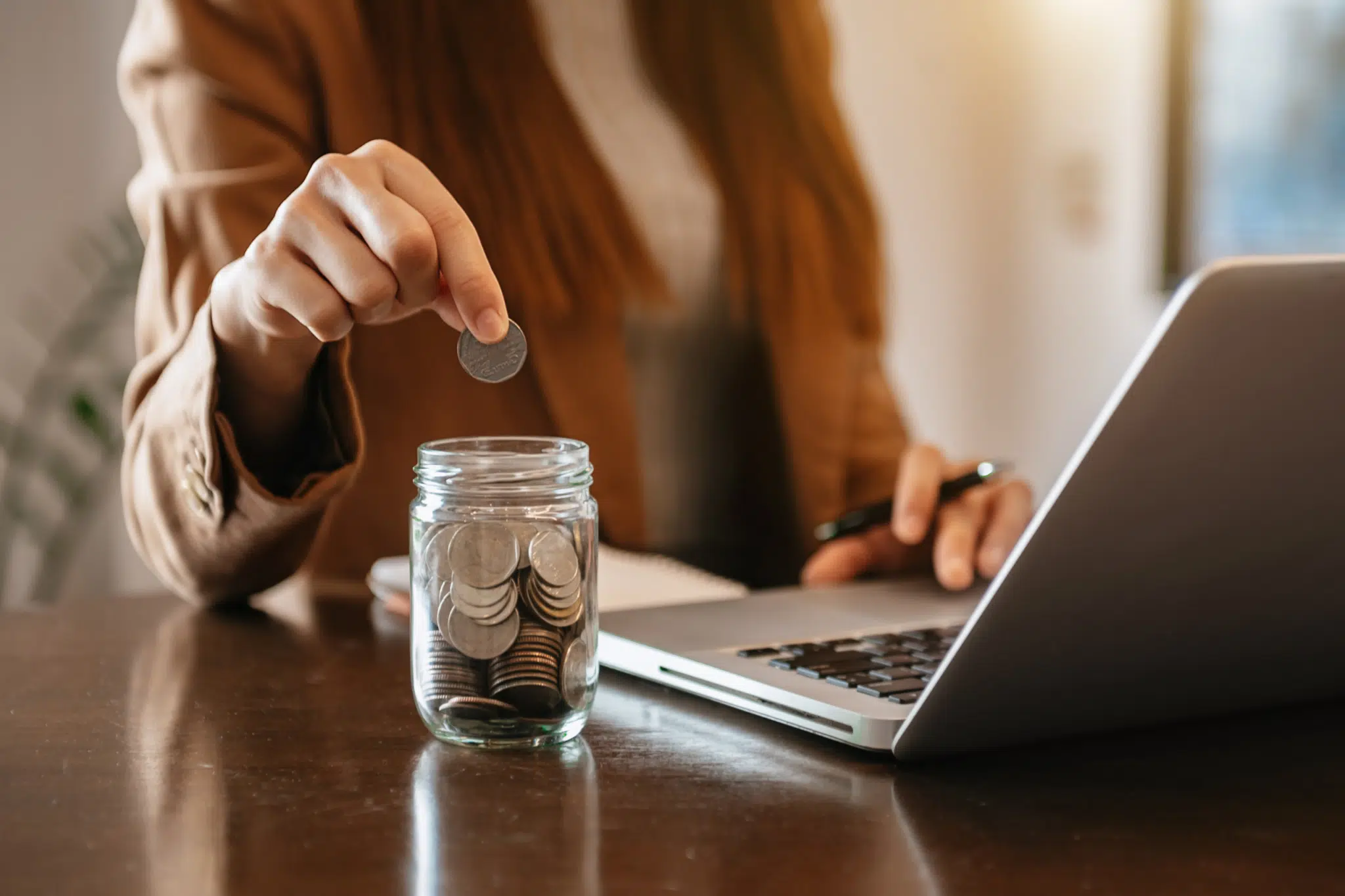 A woman puts a 50p piece in a clear jar full of other coins whilst working on a laptop